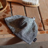 A knit sauna hat in a heathered gray and white marled pattern, resting on a rustic wooden table with a small black brand tag visible.