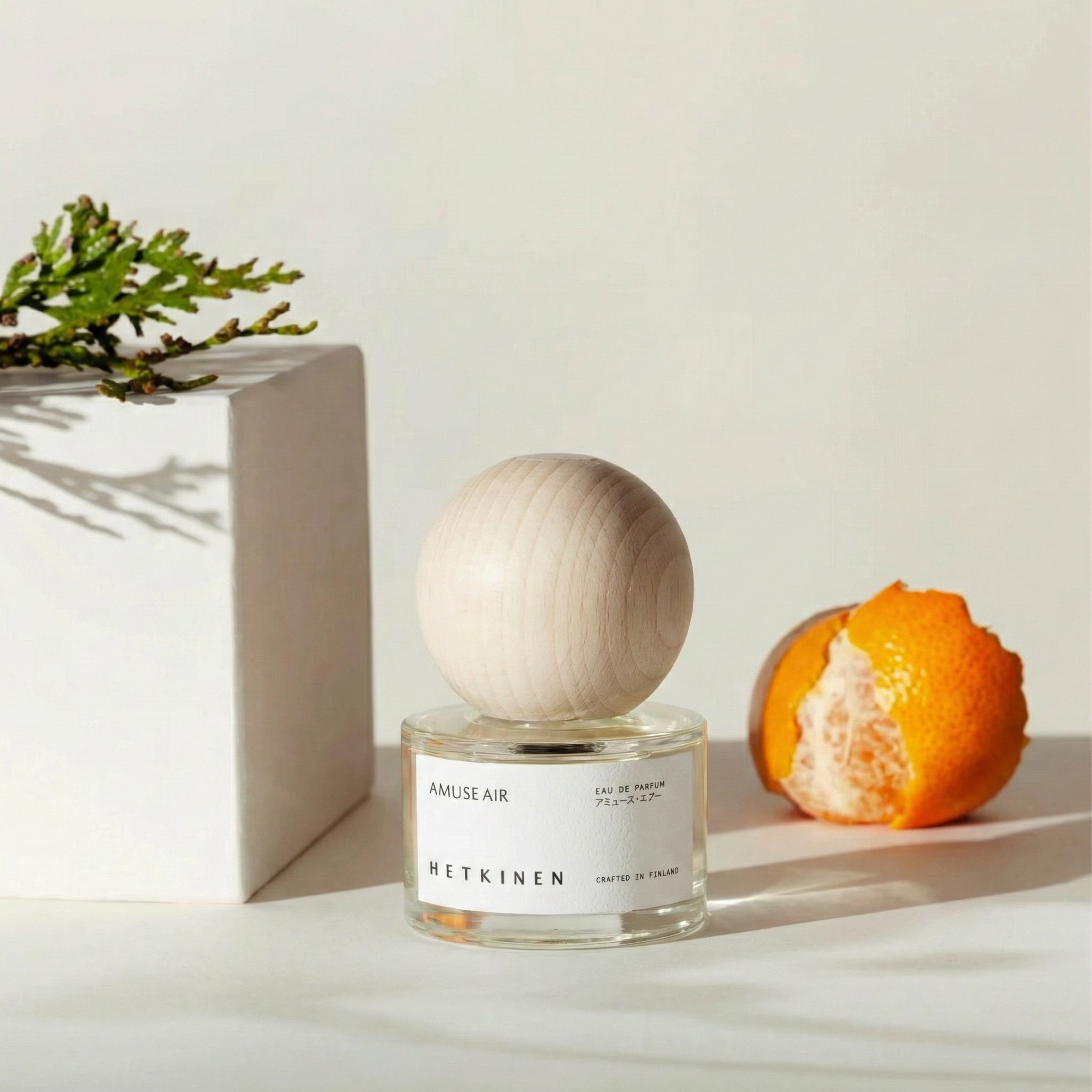 Perfume bottle with wooden cap next to an orange and plant on a light surface