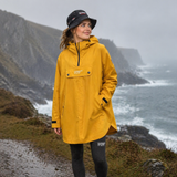 Person wearing a yellow rainponcho and navy hat standing on a coastal path with ocean and cliffs in the background.