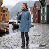 Woman in a blue raincoat standing on a cobbled street with traditional wooden houses and boats in the background.